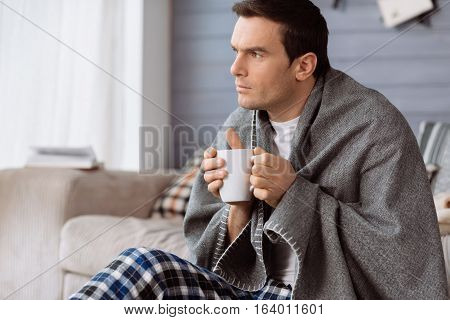 Keeping warmth. Thoughtful nice handsome man holding a cup of tea and enjoying hid drink while having a plaid on his shoulders
