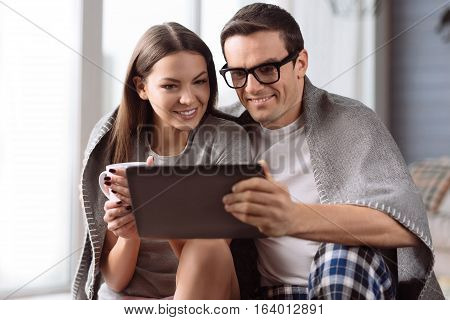 Being together. Cheerful nice young man holding a tablet and hugging his girlfriend while spending time with her