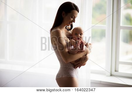 Young mother holding a naked baby on hands. She stands near the window.