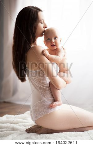 Young mother sitting with her naked baby. She holds baby in her arms. He looks with surprise.