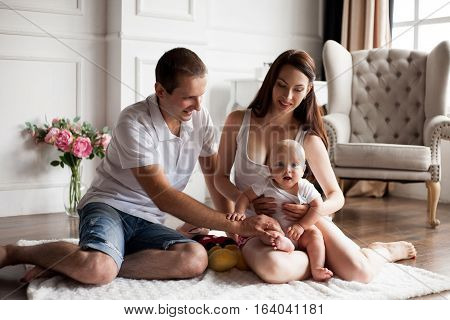 Happy young family sitting on floor in room with small baby. Beautiful interior with flowers and an armchair.