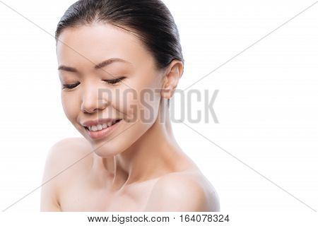 Beautiful skin. Delighted attractive Asian woman closing her eyes and smiling while standing against the white background
