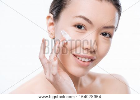Always stay young. Positive charming attractive woman putting facial cream on her cheek and smiling while caring about her face