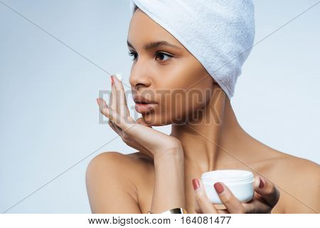 After shower. Pleasant beautiful nice woman wearing a towel turban and holding a cream bottle while applying some cream on her face