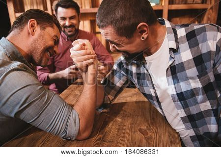 Having fun. Handsome strong pleasant man sitting opposite each other and having an armwrestling match while having fun in the pub