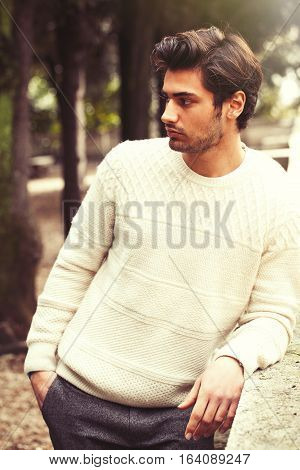 Handsome serene and pensive young man model in a trees park. Charming young and handsome man with stylish hair and white sweater. Outdoors in a park with a series of trees. Peaceful and calm attitude.