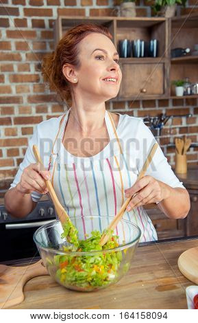 Smiling woman in apron mixing fresh vegetable salad in kitchen