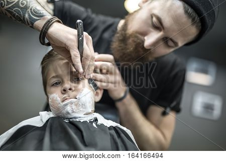 Small kid with a shaving foam on the face in the barbershop. He wears a black salon cape. Bearded barber with a tattoo is shaving boy's face with the help of the straight razor. Low aperture photo.