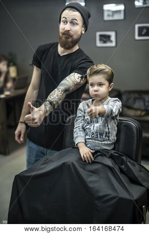 Happy small kid with the new hairstyle and the bearded tattooed barber are doing grimaces and showing the thumbs up sign. Boy wears a gray sweatshirt. Hairdresser wears a black T-shirt and cap, jeans.