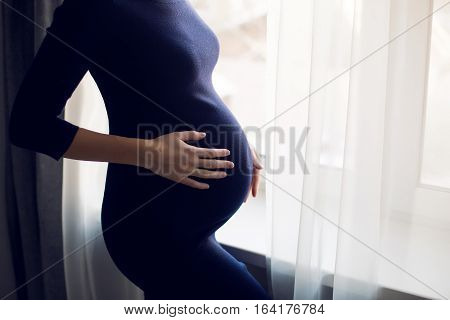 young pregnant girl in a blue bodycon dress standing at the window of the house
