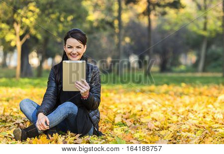 Portrait of a young happy smilling woman with a tablet sitting in a yellow autumn forest.