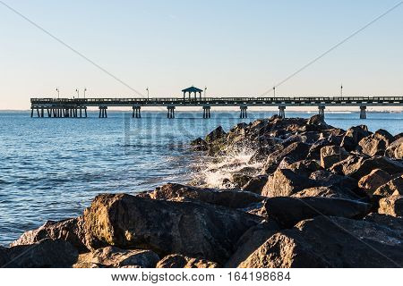 Waves crash on jetty at Buckroe Beach in Hampton, Virginia with fishing pier in background.