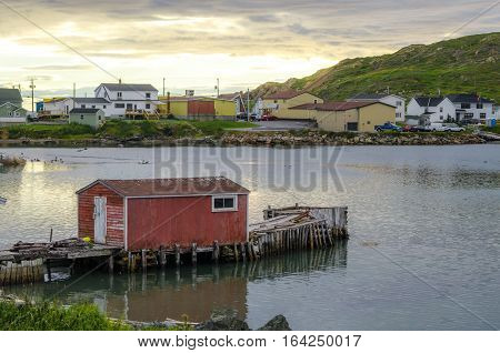 Little red shack on a dock in the small village and community of Twillingate, Newfoundland.  Sun sets brightly behind a hill in this coastal small Newfoundland community.