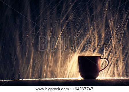 mug with warm tea in snow winter weather; backlighted cup of hot coffee on night snowy background;