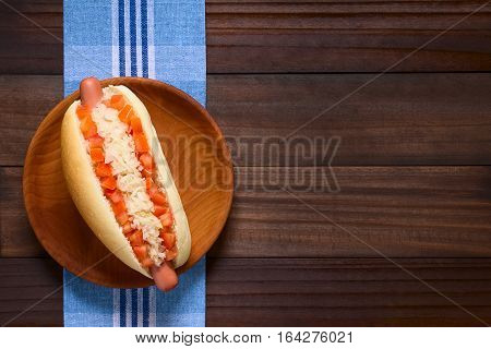 Chilean Completo Clasico (classical) or Completo Aleman (German) traditional hot dog sandwich made of bread sausage tomato cubes and sauerkraut served on wooden plate photographed overhead on dark wood with natural light