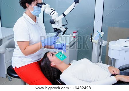 Female dentist getting ready for teeth treatment with microscope at the dental clinic. Young woman patient lying on dentist chair with open mouth. Dentist wearing mask and gloves