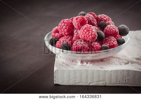 Frozen raspberries in a glass saucer. Frost on the berries. Dark background.