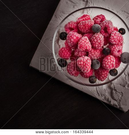 Frozen raspberries in a glass saucer. Frost on the berries. Dark background.