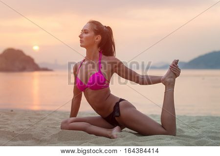 Young woman in pink bra and black panties sitting on beach stretching her legs during sunset at sea. Fitness girl doing exercises on seashore