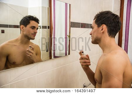Handsome shirtless young man in his home bathroom, spraying cologne or perfume on neck