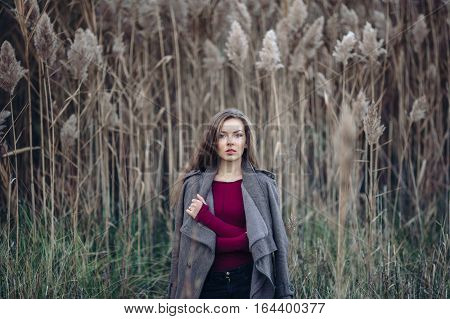 Portrait of pensive sad lonely Caucasian blonde young beautiful woman girl with long hair wearing jeans coat jacket in forest field among large tall plants grass looking in camera