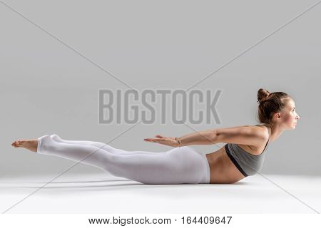 Young attractive woman practicing yoga, stretching in Double Leg Kicks exercise, Salabhasana pose, working out wearing sportswear, white pants, indoor full length, isolated, grey studio background