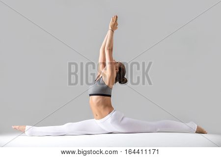 Young attractive woman practicing yoga, sitting in Monkey God, Splits exercise, Hanumanasana pose, working out wearing sportswear, indoor full length, isolated against grey studio background