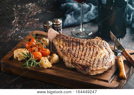 Succulent grilled tomahawk beef steak on the bone with red wine, seasonings, fresh rosemary and grilled vegetables on cutting board