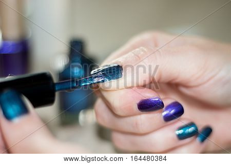 hands of a young woman who is painting her nails with metalized dark green and dark blue nail varnish