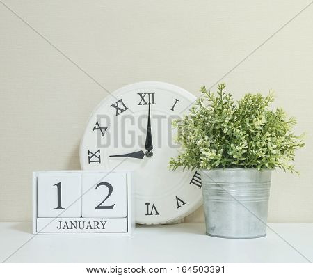 White wooden calendar with black 12 january word with clock and plant on white wood desk and cream wallpaper textured background selective focus at the calendar