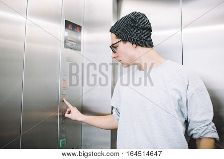 Young stylish guy standing in elevator. Attractive boy in hat and glasses presses button to go up for the meeting. View in profile