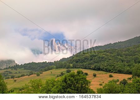 View from far away of rocks of the Picos de Europa National Park. In a gap between clouds peaks of mountains in the foreground the field and the wood are visible