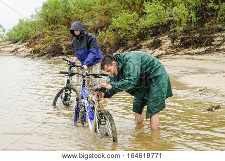 Abalak, Russia - August 11, 2010: Two dirty cycle tourists wash bicycles from dirt during rain in Irtysh river. Stop on a lodging for the night near the village