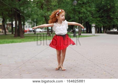 Little girl with red hair dances in green summer park shallow dof