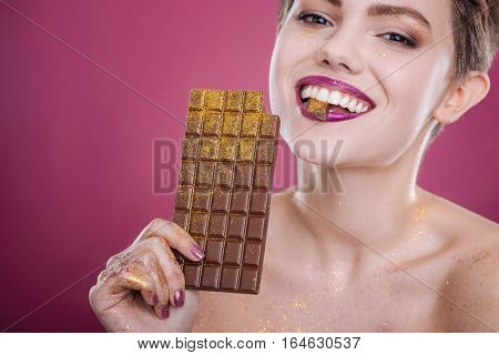 So delicious. Cheerful delighted young woman holding chocolae bar covered with tinsel while smiling