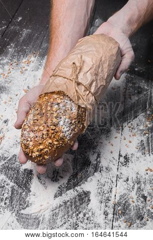 Warm fresh rye whole grain bread, wrapped in craft paper. Healthy baking and cooking concept background. Hands carefully hold loaf on wooden table background, sprinkled with flour. Soft toning