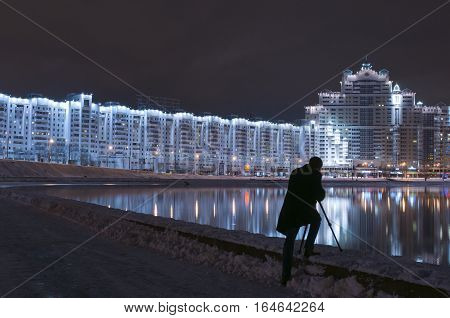 Man shooting reflections in river. Silhouette of man photographer with digital camera and tripod taking picture of night view of city in winter.