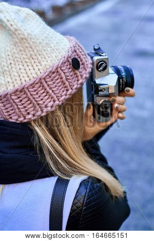 Beautiful girl photographer photographing the old town with retro camera. She is wearing a knitted cap