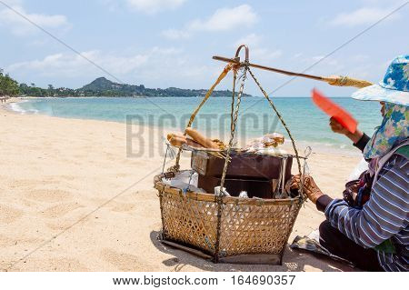 Unidentifiable vendor with a basket on a sandy beach in Thailand. The person is grilling sausages.
