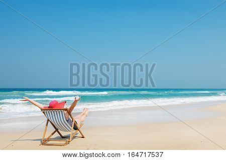 Happy woman in red sunhat on the beach sitting on deckchair with hands up.Vacation and travel concept