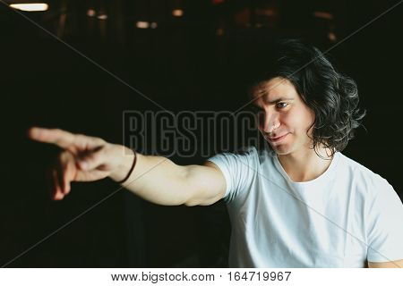 Beautiful long-haired brunette smiling man in a white T-shirt It shows his hand towards