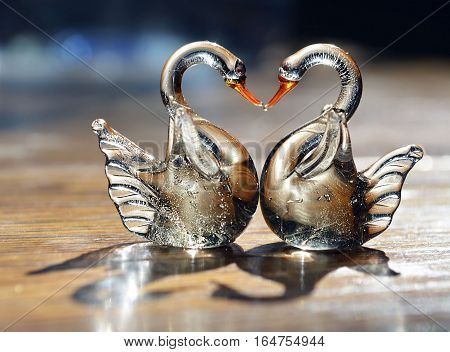 Two glass swans with necks forming a love heart shape.Figurine glass swans on wooden background.Saint Valentine background with two swans in love.Valentines day or love concept.Soft selective focus.