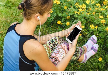 close up woman in sportswear typing on mobile phone in hands with white headphones sitting in the grass on the background of nature