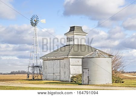 A small white barn and windmill stand under a cloudy blue Midwestern Sky.