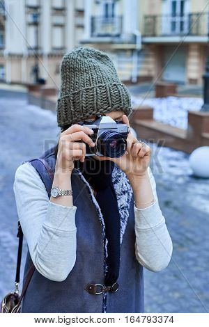 Beautiful girl photographer photographing the old town with retro camera. She is wearing a knitted cap