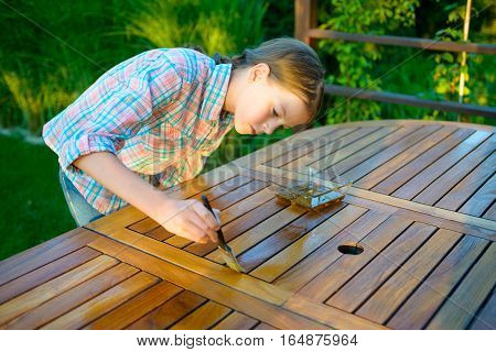 Young pretty girl holding a brush applying varnish paint on a wooden garden table