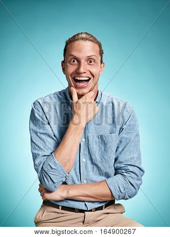 Happy excite young man in blue shirt smiling and celebrating his succcess over blue studio background