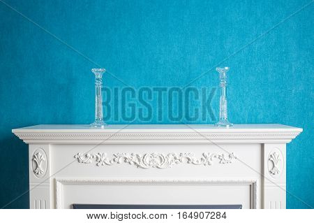 Crystal candlesticks stand on a white fireplace with a decor. The blue wall acts as a background.