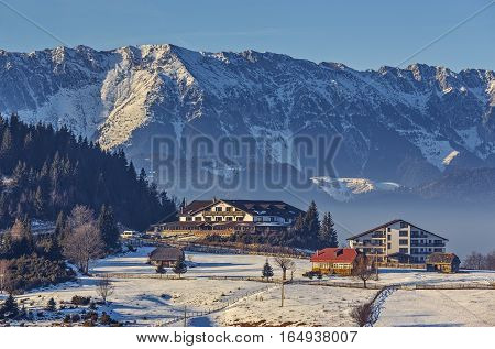 Sunny winter scenery with majestic snowy Piatra Craiului mountains chalet or lodging buildings in Cheile Gradistei Fundata touristic resort Romania. Idyllic quiet holiday retreat destinations.