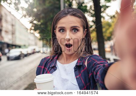 Attractive young brunette woman with a shocked expression talking a selfie while out walking in the city